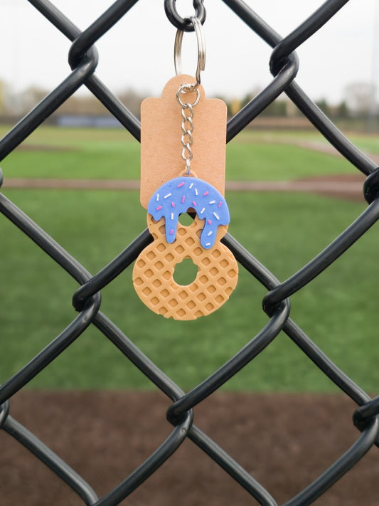 A 3D printed keychain shaped like a drippy ice cream cone with baseball number 8, attached to a chain on a fence, with a grassy field in the background.