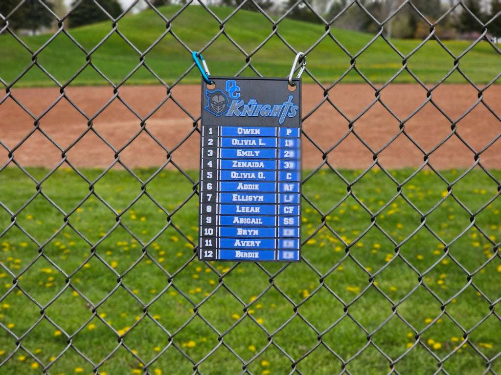 Scoreboard with player names and numbers attached to a chain-link fence on a baseball field.