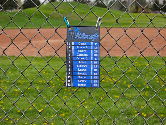 Scoreboard with player names and numbers attached to a chain-link fence on a baseball field.